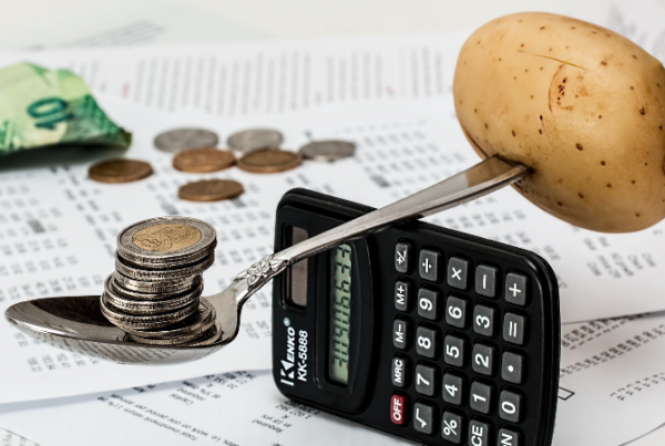 A spoonful of coins with a potato pierced into the other end, balancing on a calculator, depicting a precarious balance of budget.