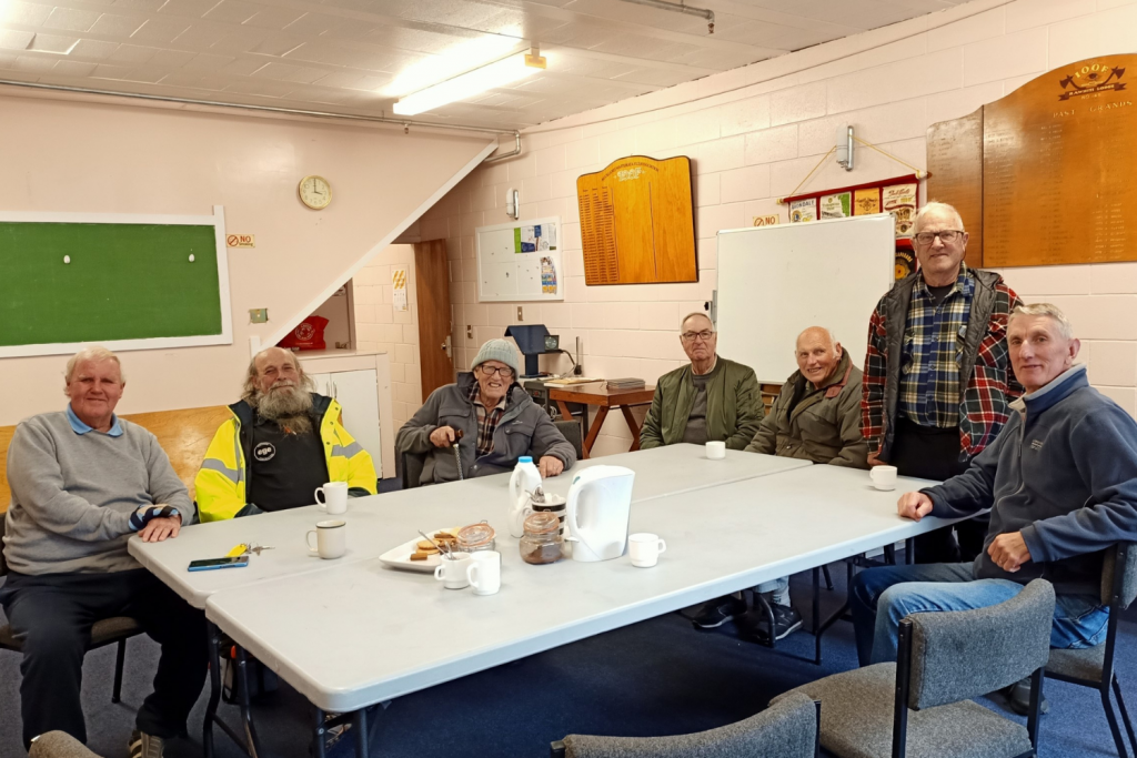 A group of seven older men sat around a table enjoying tea, biscuits and companionship.