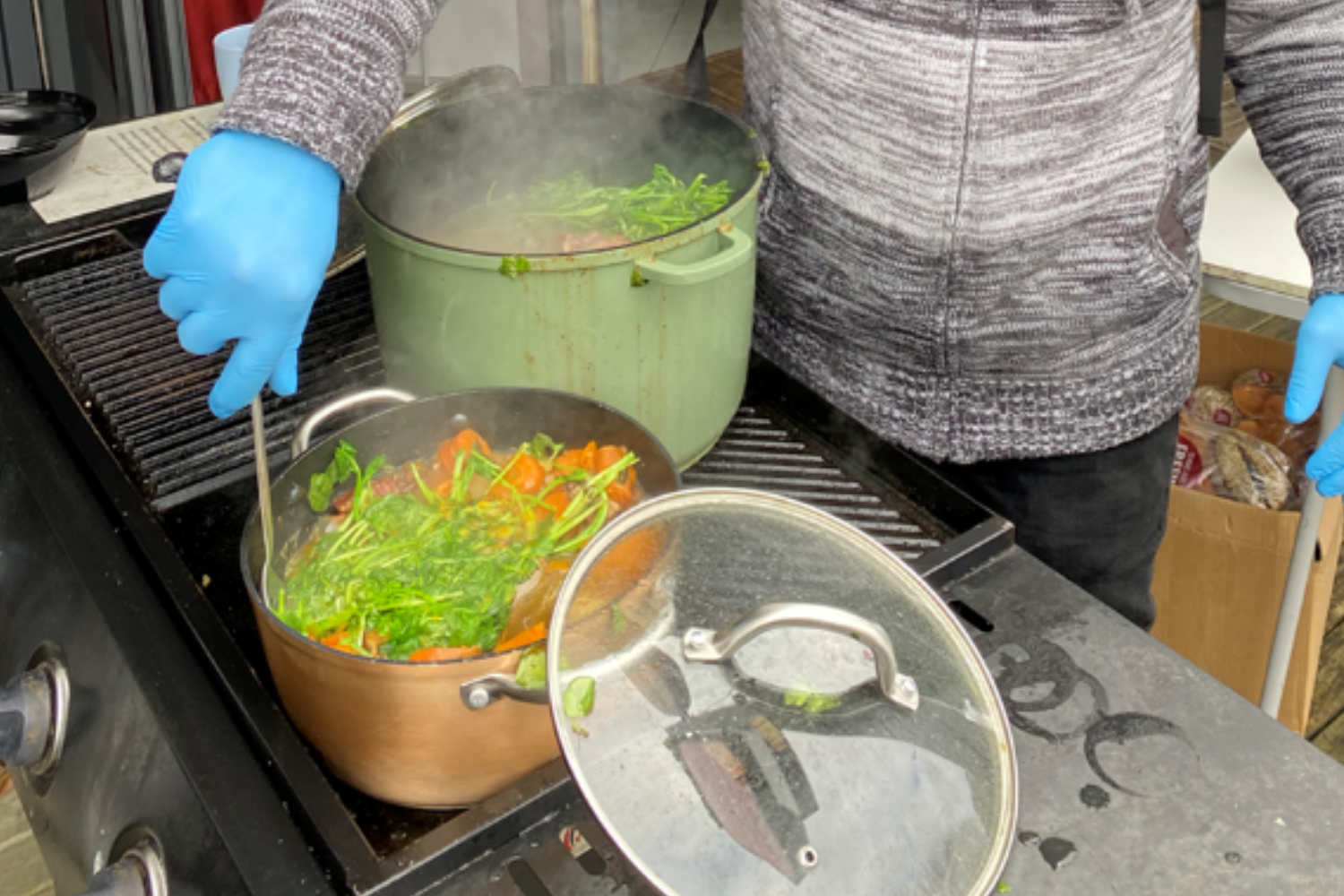 Close-up of a boiling pot of food, man wearing blue gloves stirring the vegetables and boil-up ingredients.