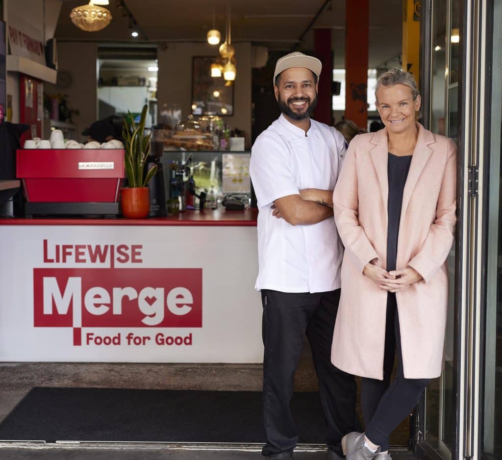 Two smiling individuals, a chef in white attire and a customer in a pink coat, standing proudly in front of a café named "Lifewise Merge - Food for Good."