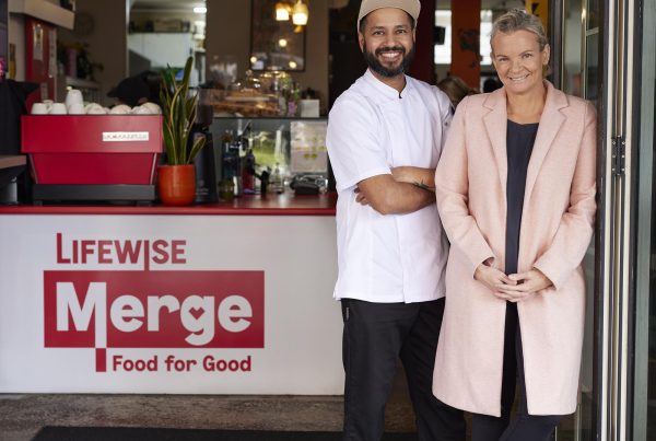 Two smiling individuals, a chef in white attire and a customer in a pink coat, standing proudly in front of a café named "Lifewise Merge - Food for Good."